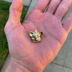 A24 Uncut Gems Gold with blue dots furby enamel pin held in a hand to show small size with a blurred outdoor background