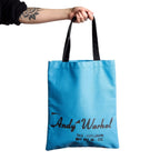 Blue tote bag with 'Andy Warhol' text held by a person on a white background