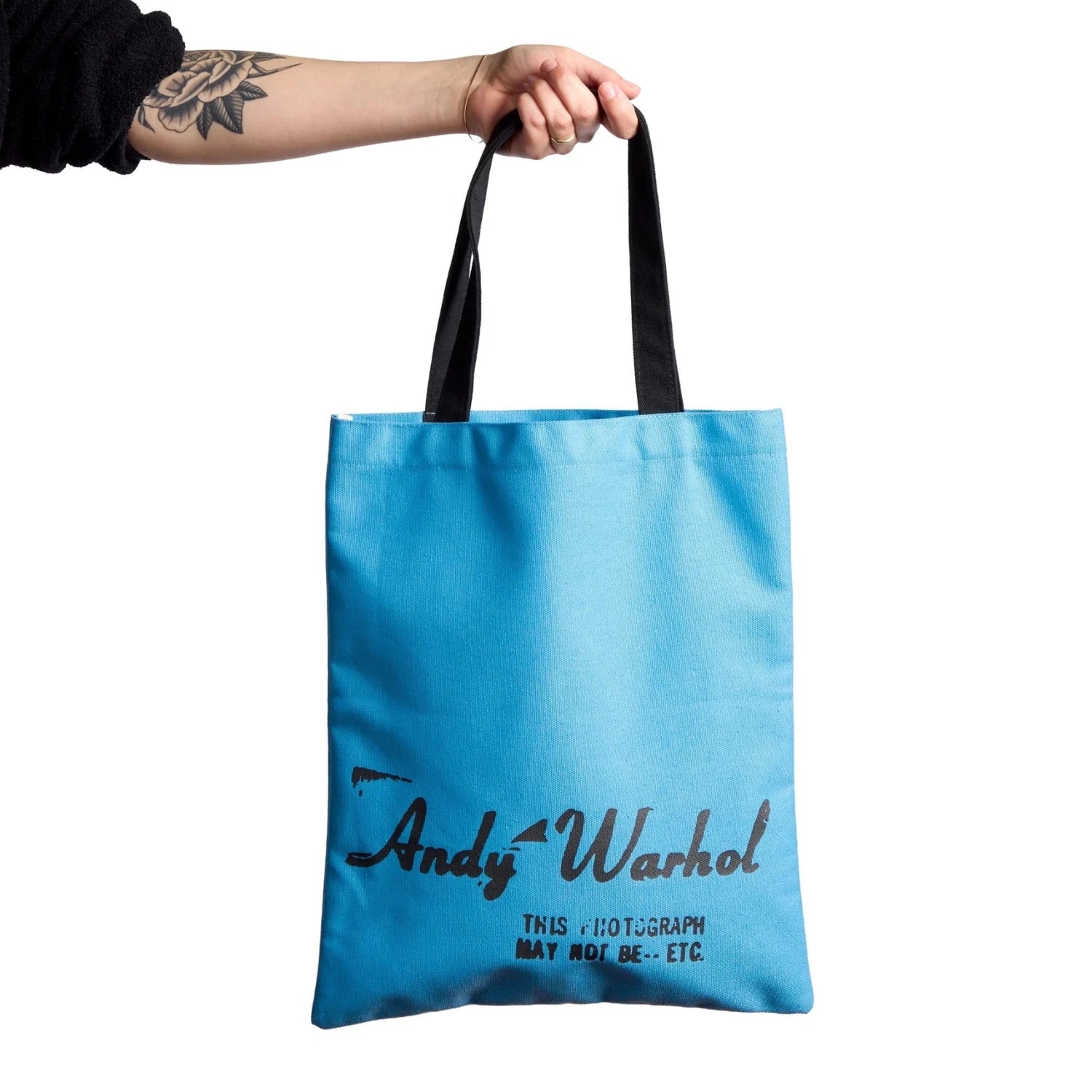 Blue tote bag with 'Andy Warhol' text held by a person on a white background