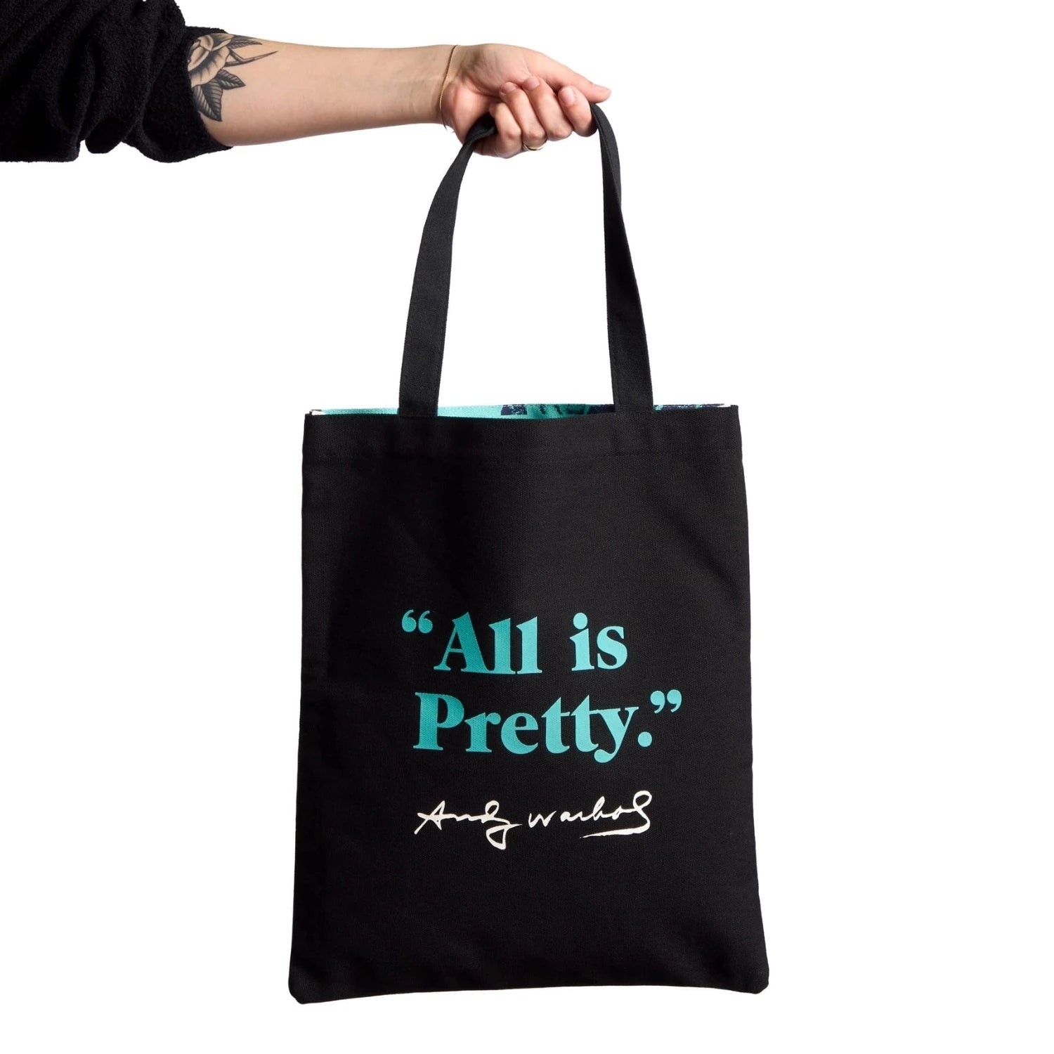 Black tote bag with 'All is Pretty - Andy Warhol' text held by a person on a white background