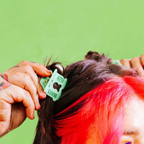 Person with colorful hair putting a Green plastic hair claw clip that looks like a bread clip in her hair against a green background