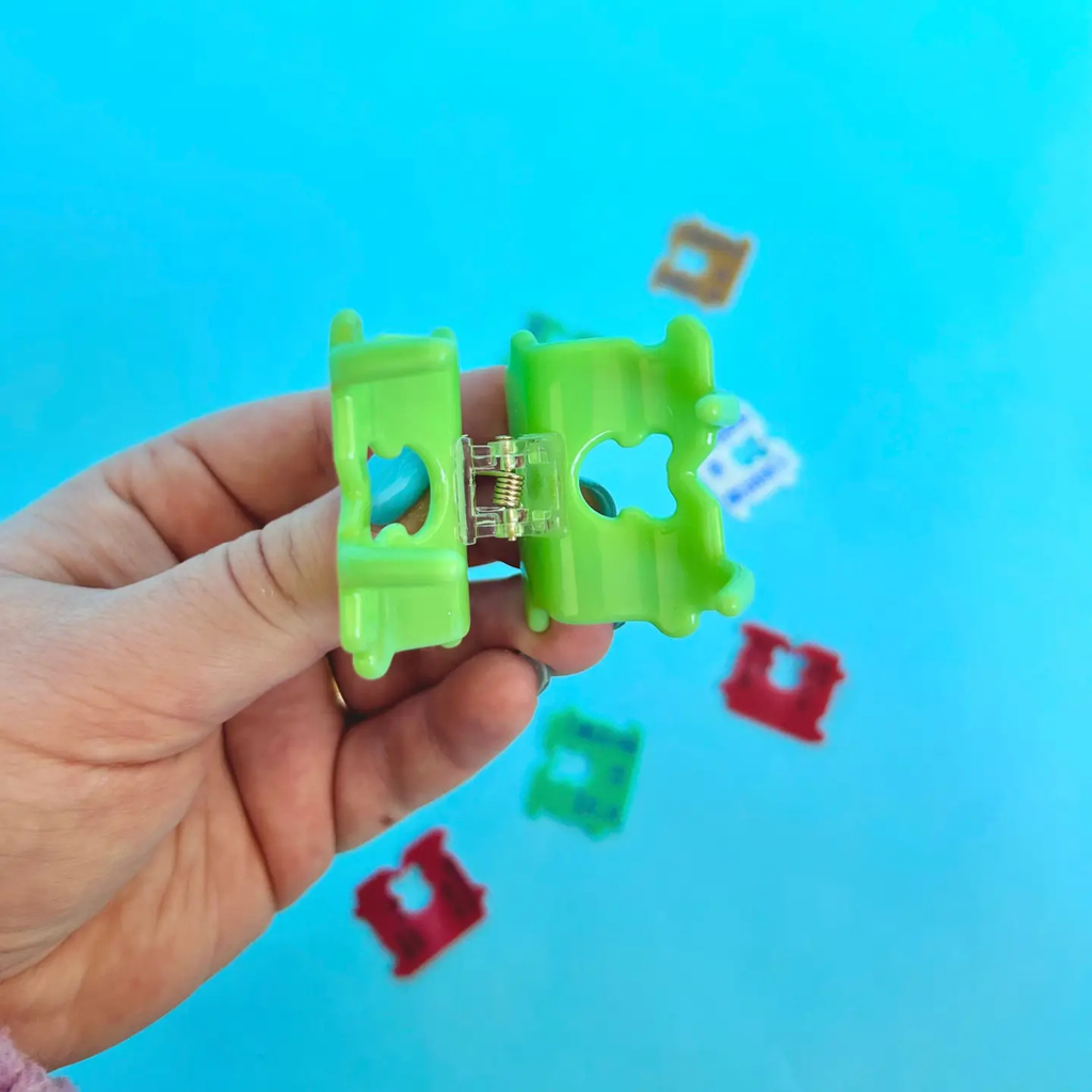 Hand holding a green plastic clip against a blue background with colorful clips scattered around.