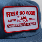 Blue chambray trucker cap with a 'Feels So Good' trucker style patch featuring text and graphics.
