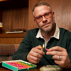 Seth Rogen lighting a colorful incense with a lighter in a warm-toned room.