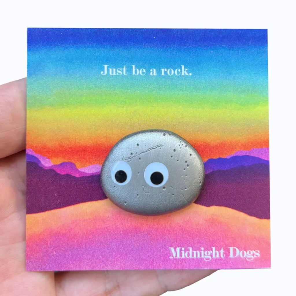 Pin of A24's Everything Everywhere All At Once rock design and googly eyes on a colorful card with 'Just be a rock' text, held by a hand.