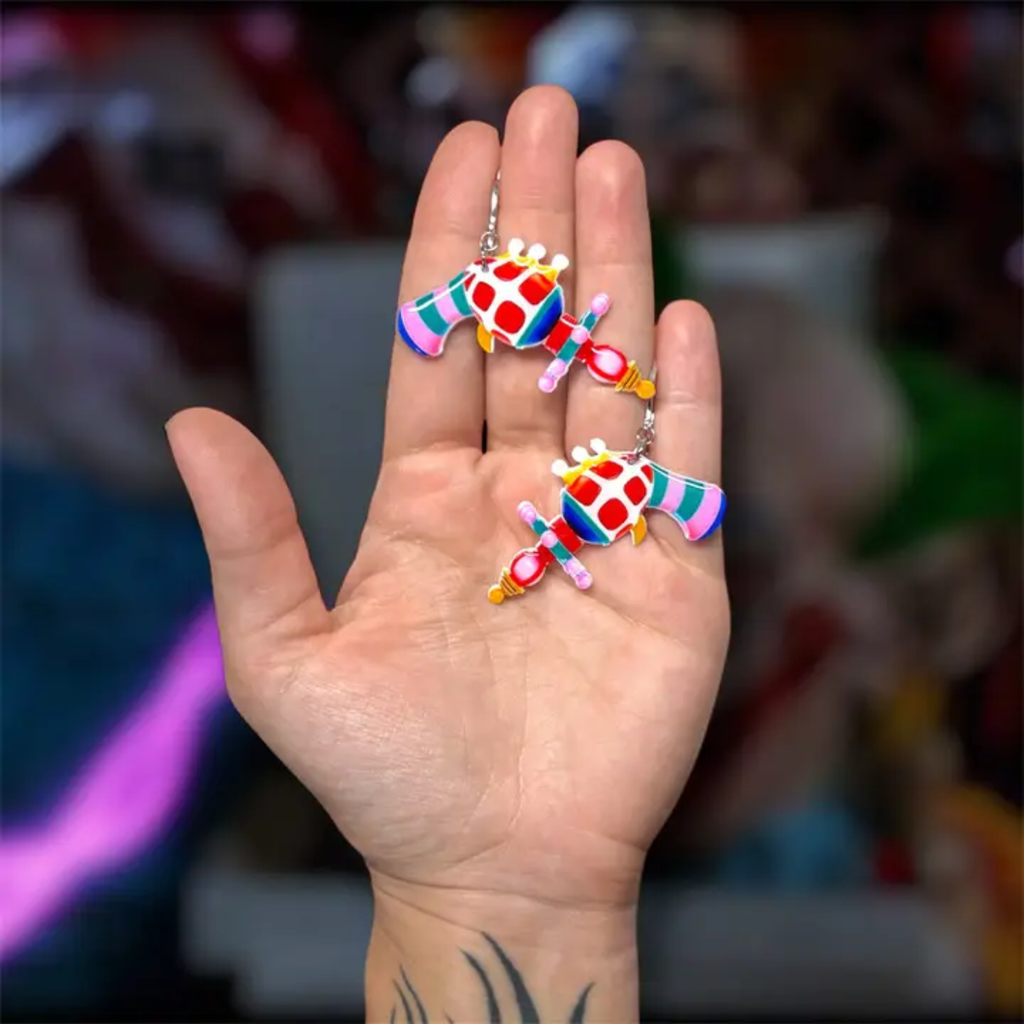 A hand holding two colorful, handcrafted earrings shaped like circus ray guns. The earrings feature a mix of pink, blue, green, yellow, and red colors, shown against a blurred background.