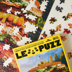 Jigsaw puzzle with a partially completed design of a sandwich, featuring the 'Le Puzz' packaging.