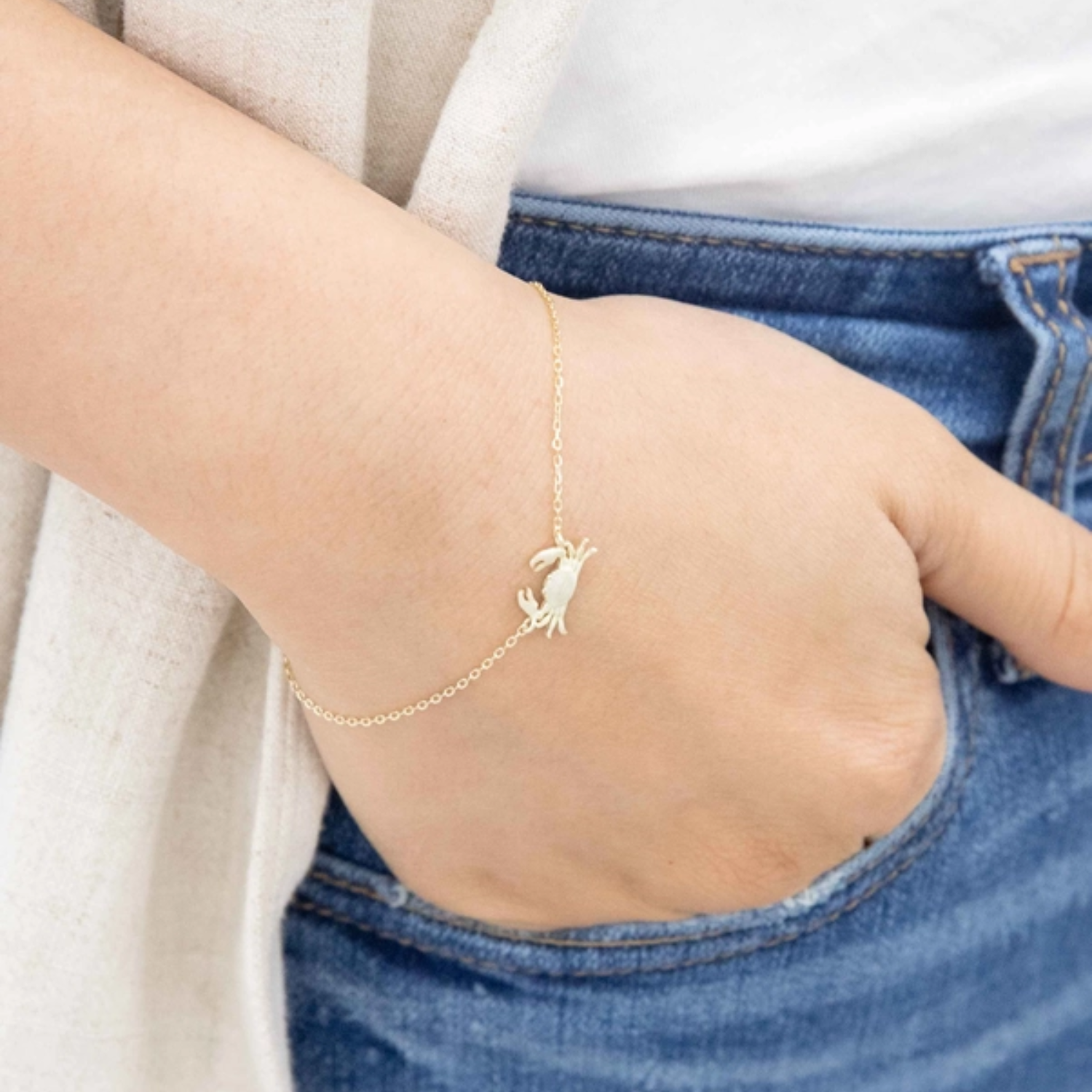 A delicate gold chain bracelet with a small Maryland Blue Crab charm worn on a person's wrist, shown in a close-up lifestyle shot with blue jeans and a white top.