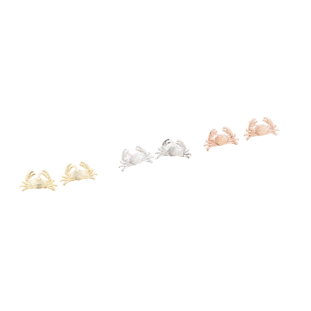 Three pairs of small crab-shaped stud earrings in yellow gold, silver, and rose gold tones, arranged horizontally on a plain white background.