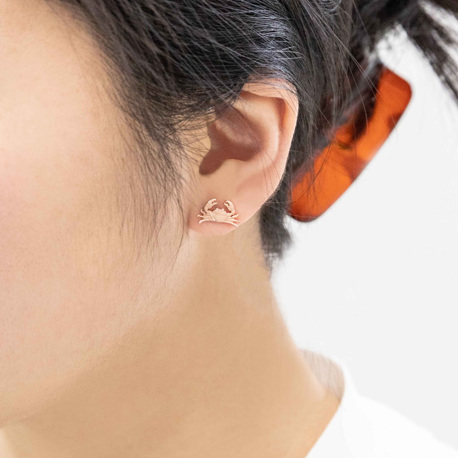 A close-up of a person's ear wearing a small rose gold crab stud earring, with dark hair and an orange hair tie visible against a white background.