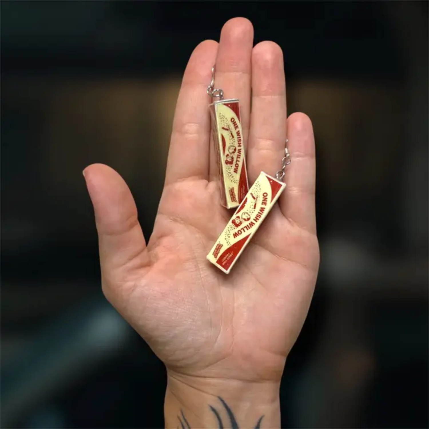 A hand holding two small rectangular pendant earrings with silver-toned chains. The pendants resemble One Wish Willow boxes from the horror movie 'Obsessed'.