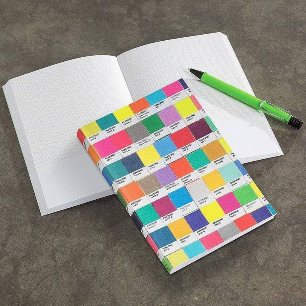 Pantone Color swatch notebook with colorful squares on a gray surface with an open notebook and green pen.