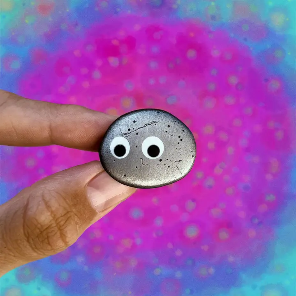Hand holding Everything Everywhere All At Once pin of rock with googly eyes against a colorful, abstract background