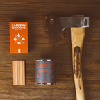 Camping-themed items including a campfire cologne box, candle, and axe on a wooden surface.