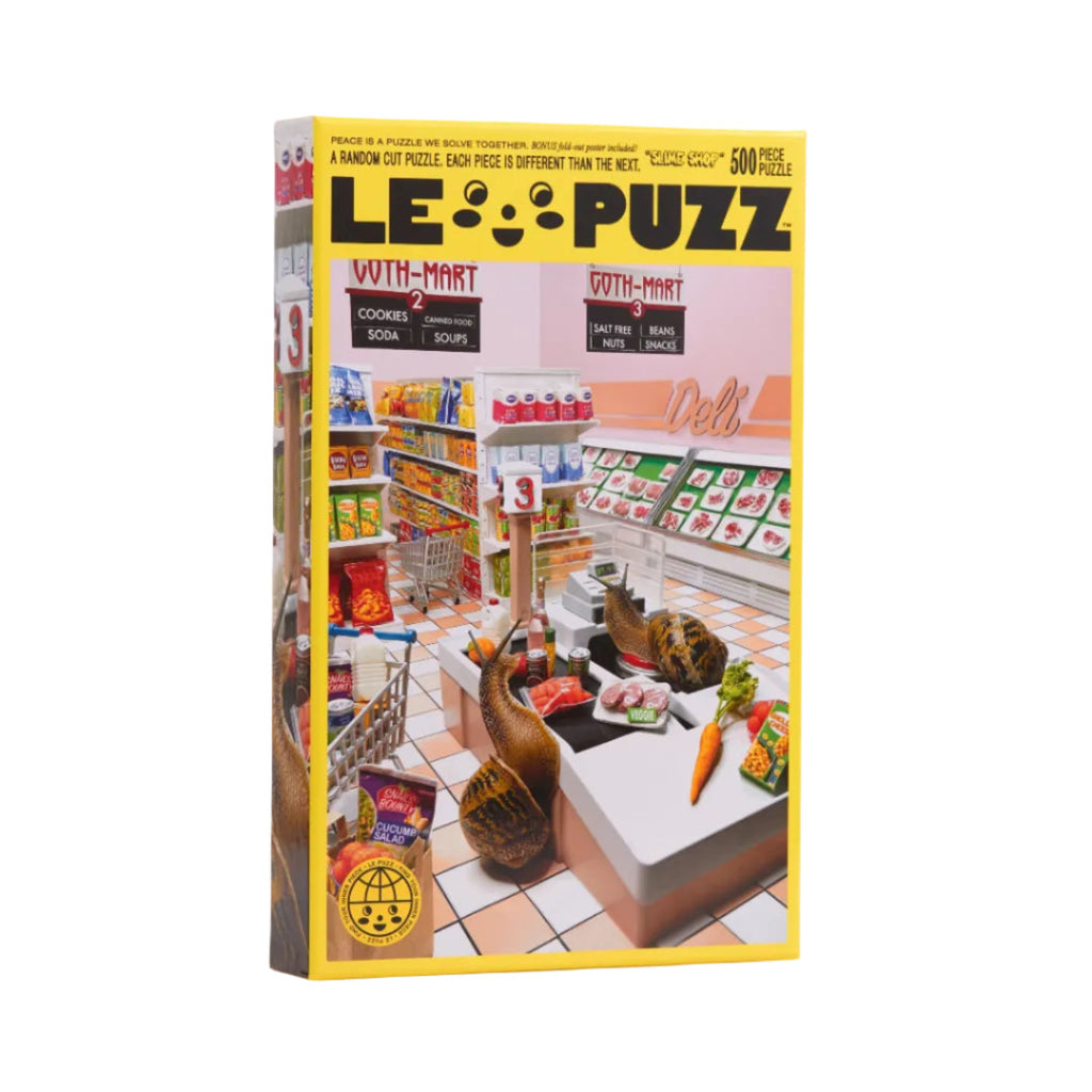 A boxed jigsaw puzzle standing upright on a white surface against a light gray background. The box is labeled 'LE PUZZ' and shows a colorful illustration of a supermarket interior with shelves, checkout counters, and produce.