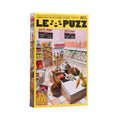 A boxed jigsaw puzzle standing upright on a white surface against a light gray background. The box is labeled 'LE PUZZ' and shows a colorful illustration of a supermarket interior with shelves, checkout counters, and produce.