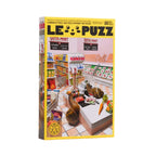 A boxed jigsaw puzzle standing upright on a white surface against a light gray background. The box is labeled 'LE PUZZ' and shows a colorful illustration of a supermarket interior with shelves, checkout counters, and produce.