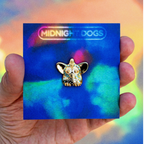 A24 Uncut Gems Gold with blue dots furby enamel pin on a white background 'Midnight Dogs' card held by a hand against a colorful background