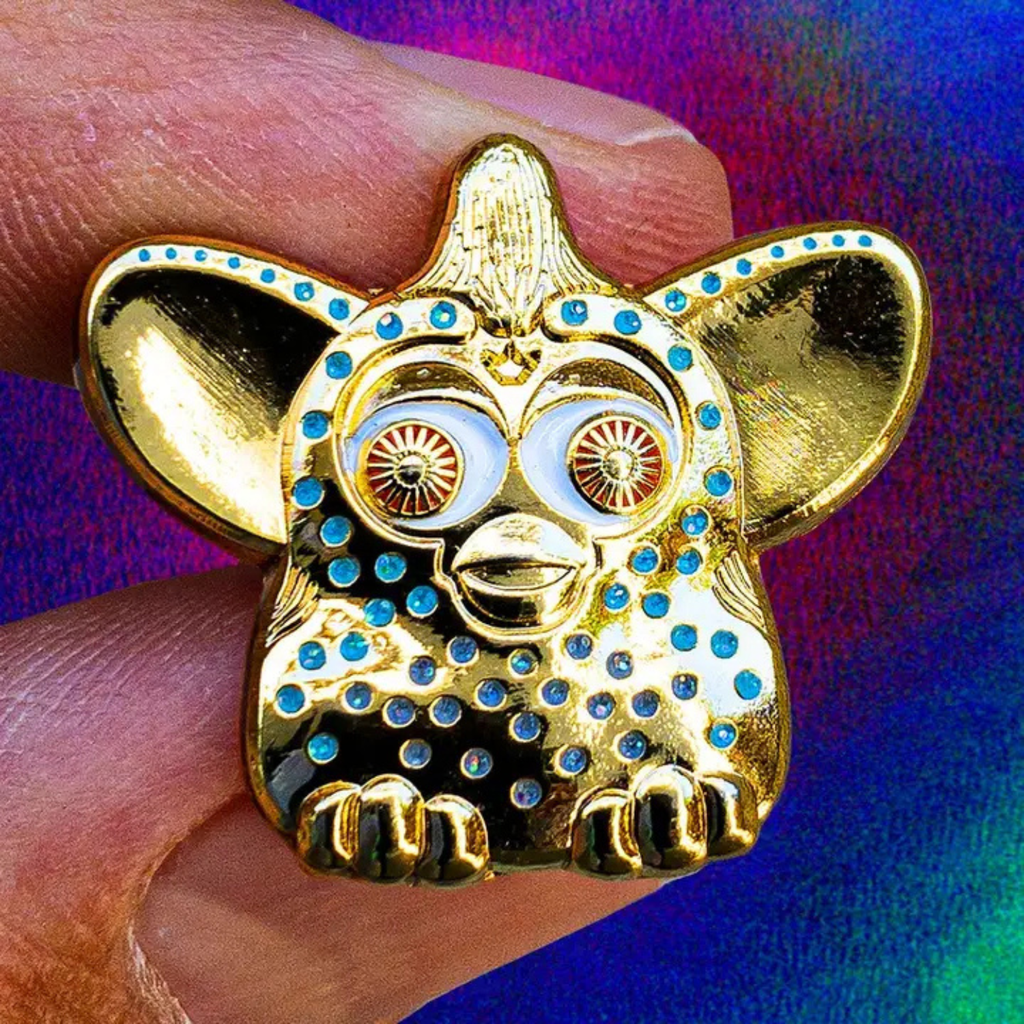 A24 Uncut Gems Gold with blue dots furby enamel pin held between fingers against a colorful background