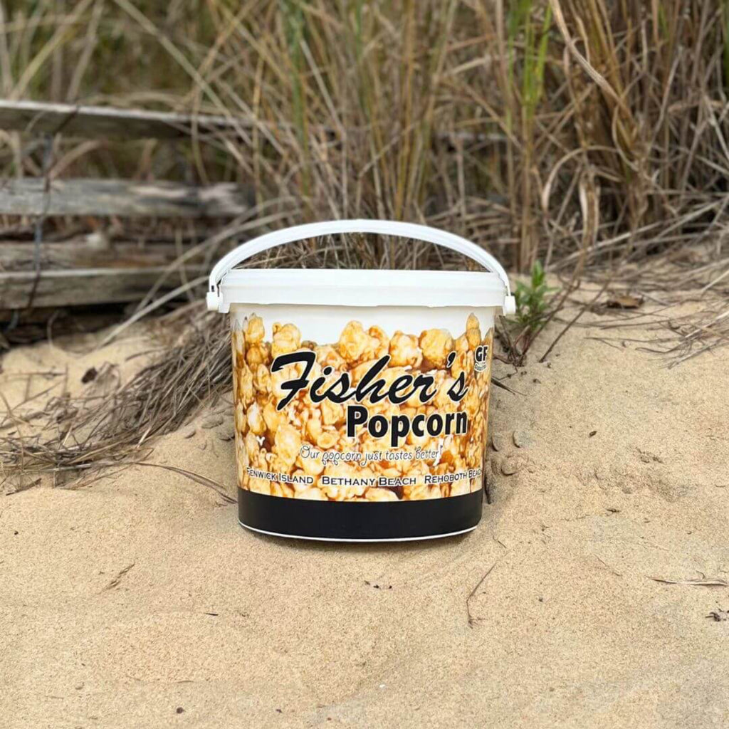 Fisher's Popcorn bucket on a sandy beach with grass and driftwood in the background