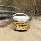 Bucket of Fisher's Popcorn on a sandy beach with grass and driftwood in the background.