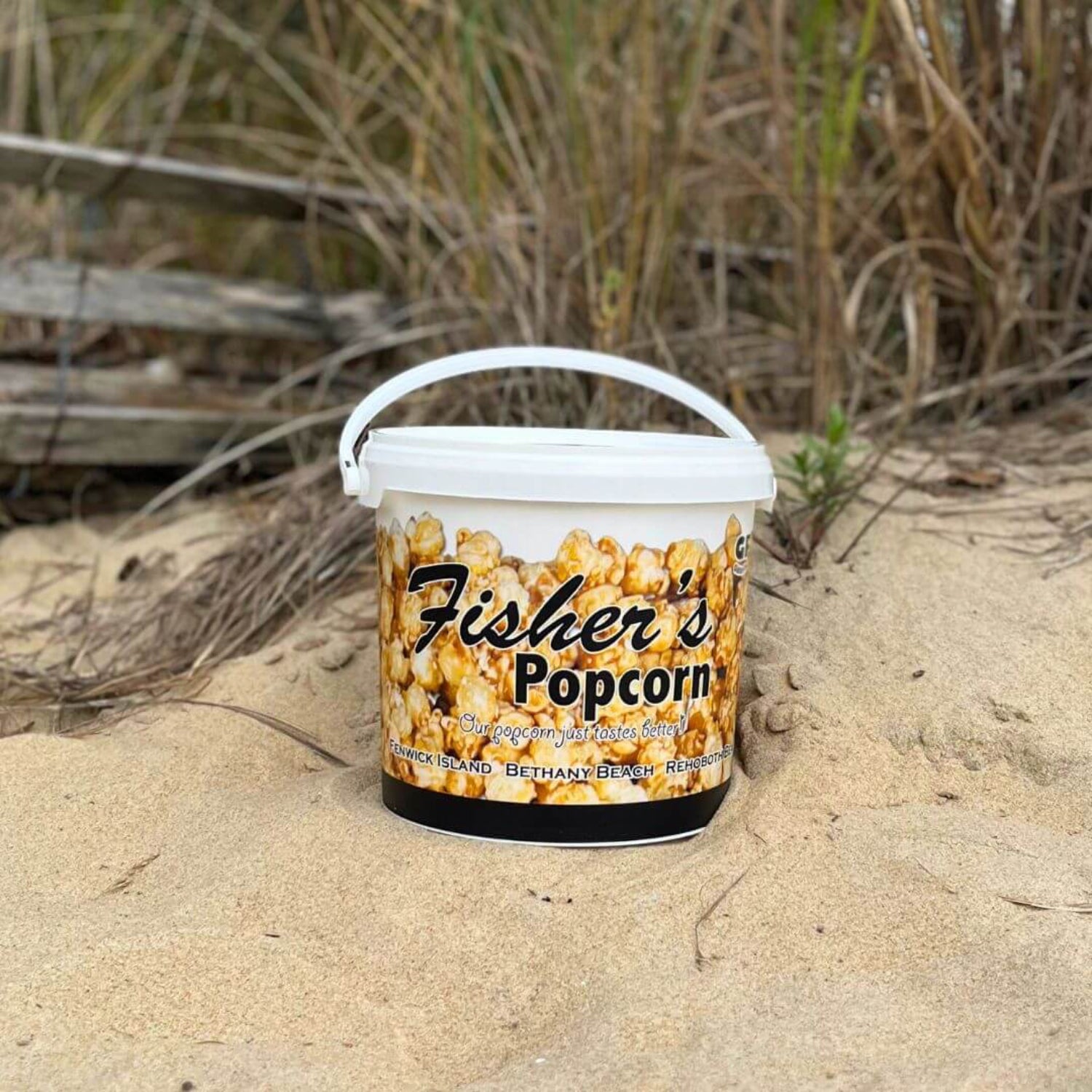 Bucket of Fisher's Popcorn on a sandy beach with grass and driftwood in the background.